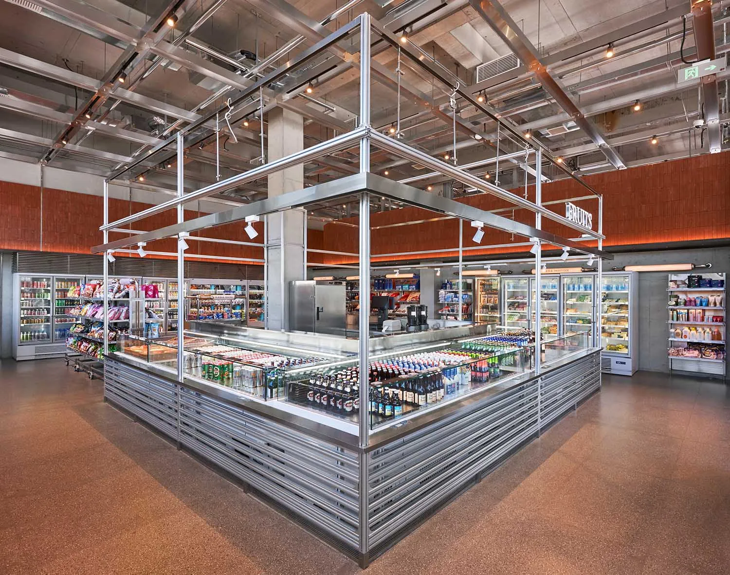 Wide view of a modern supermarket featuring a large stainless steel central display island filled with beverages, surrounded by refrigerated cases and industrial metal ceiling structure.