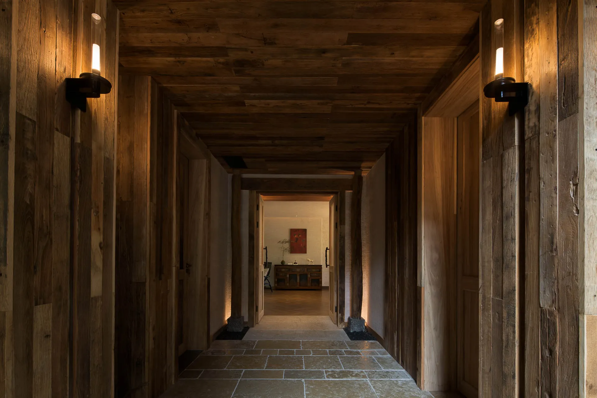 Long rustic hallway completely lined with reclaimed wood planks on walls, ceiling and doors, featuring black modern wall sconces casting warm light on the stone-tiled floor.
