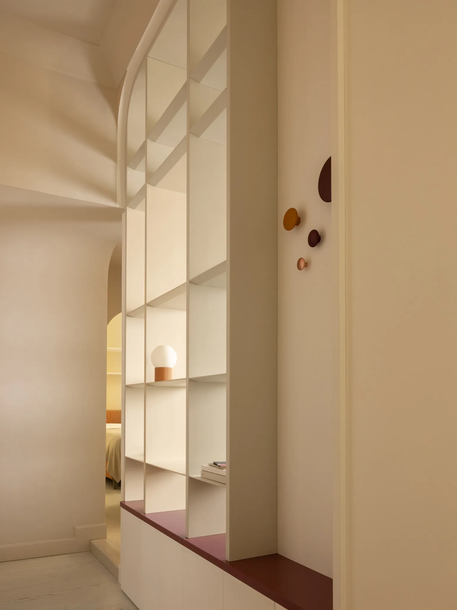 Modern arched built-in shelving unit in off-white with terracotta-colored base and decorative hooks, featuring a spherical table lamp on one shelf.