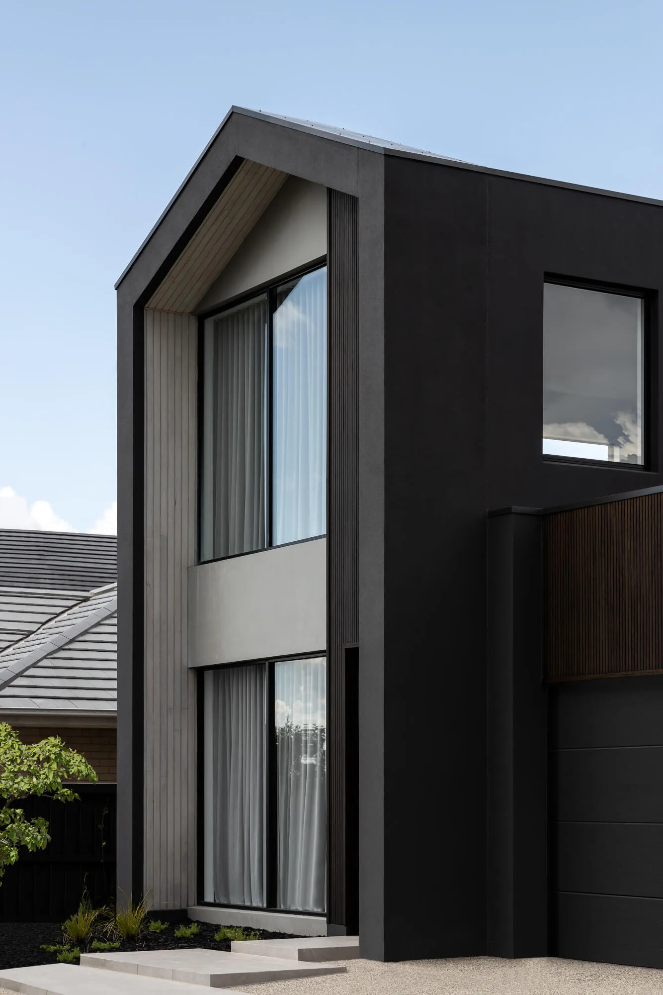 Modern two-story house exterior featuring charcoal black cladding, vertical light timber slats, large double-height windows with sheer curtains, and concrete steps under a clear blue sky.