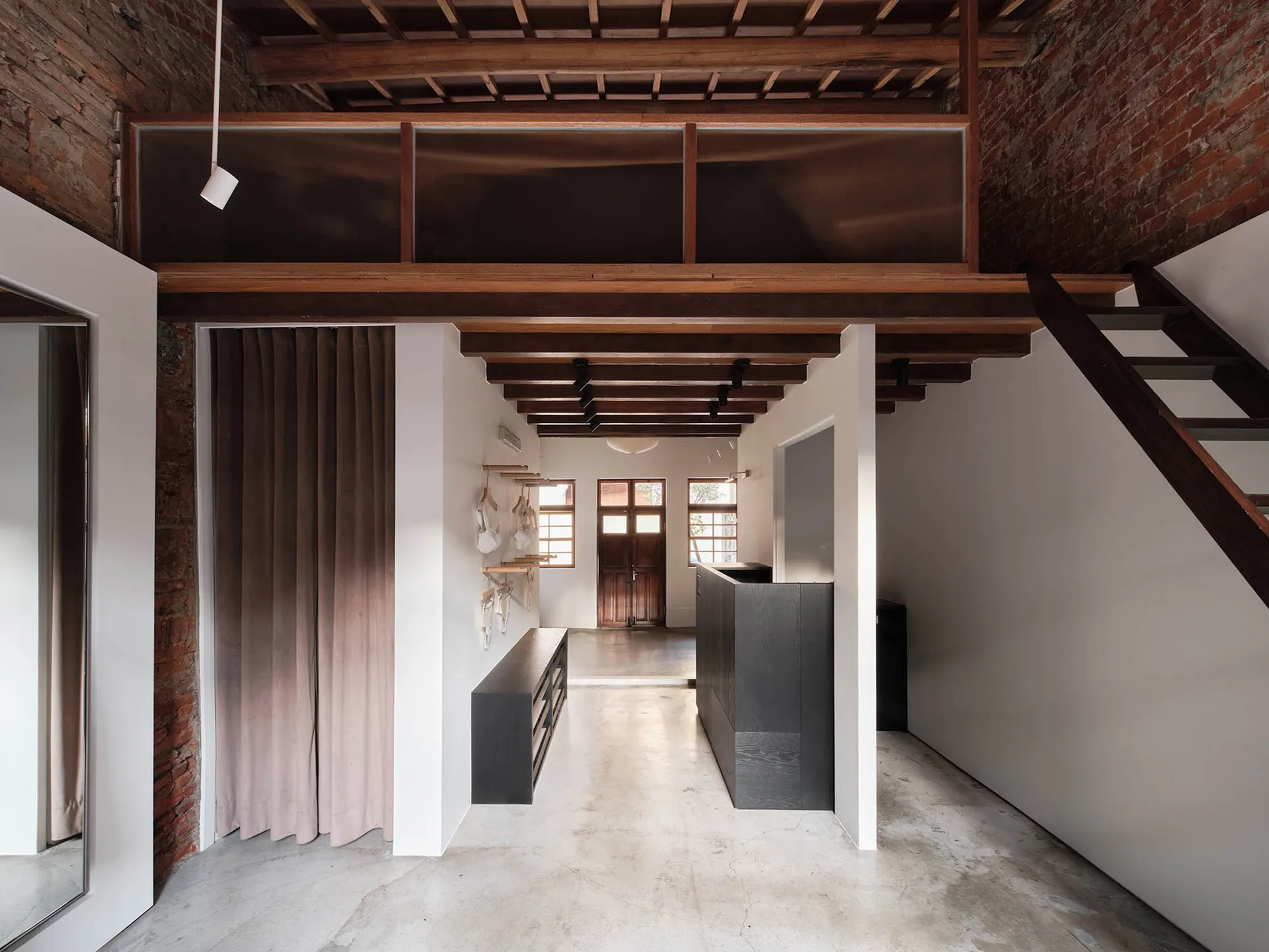 View of a modern industrial loft entryway featuring exposed red brick walls, heavy wooden ceiling beams, a dark wood staircase on the right, and a full-height pink velvet curtain on the left.