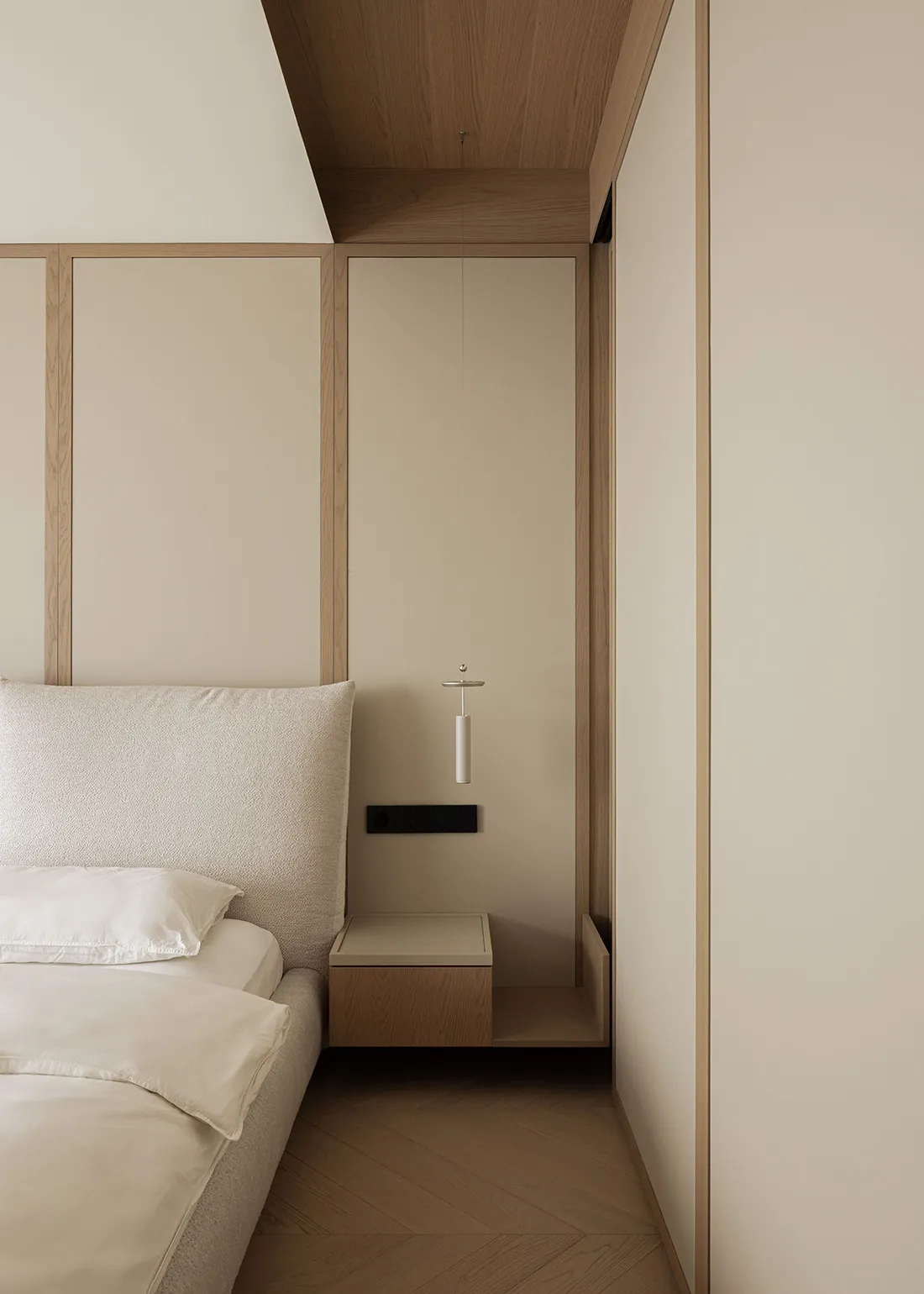 Modern minimalist bedroom corner showing a beige upholstered bed with white linens, a wall-mounted oak floating nightstand, and a slim white pendant light hanging from a wood-paneled ceiling.