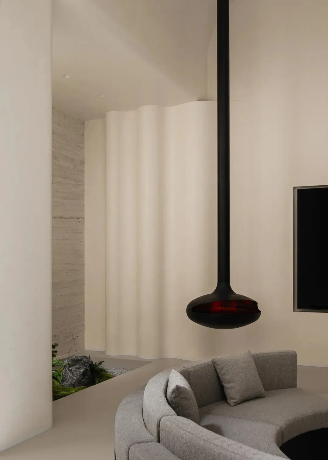 Modern living room featuring a curved grey fabric sofa, a large black sculptural pendant lamp, a textured travertine wall, and an indoor rock garden with moss and ferns.