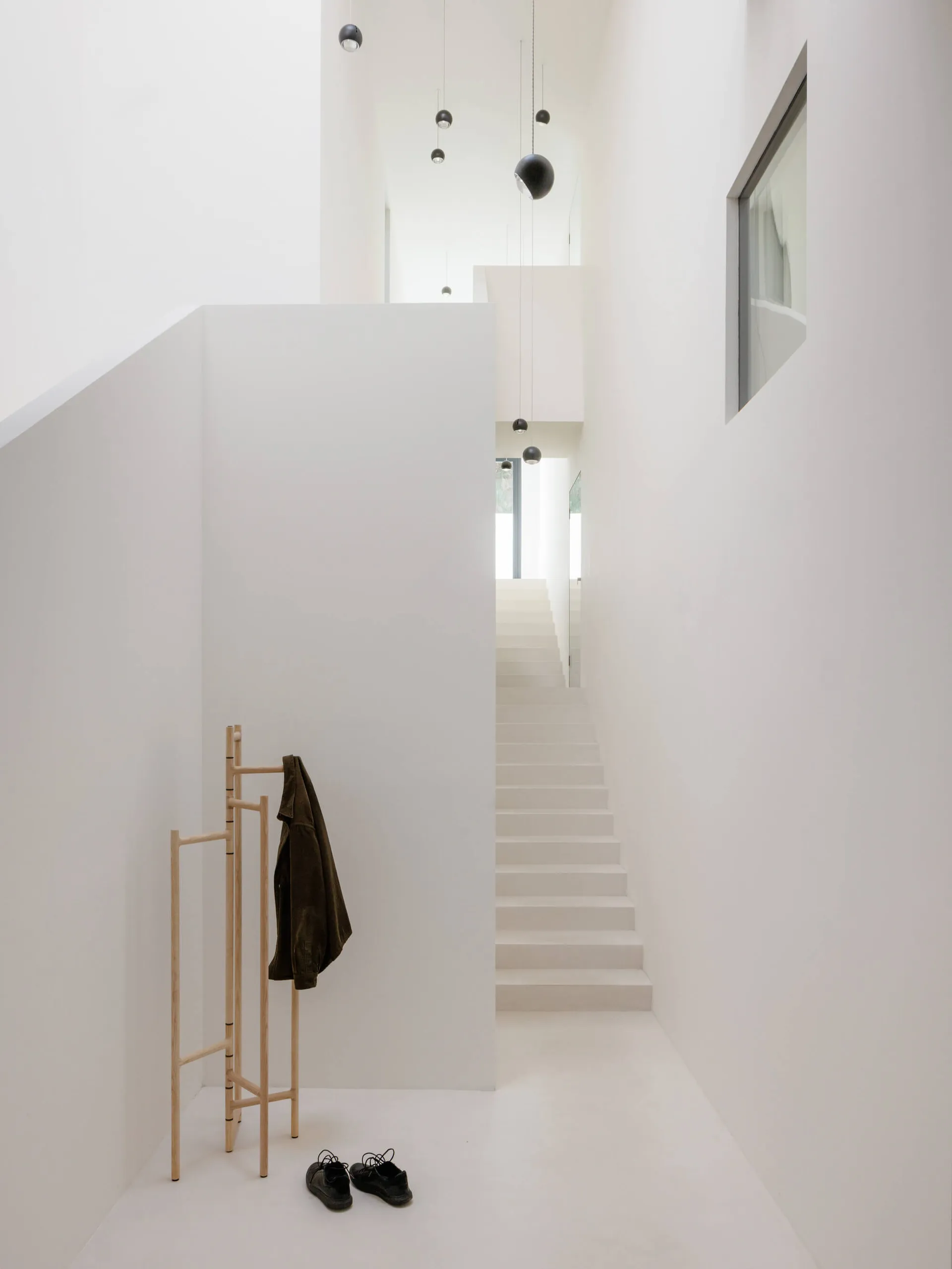 Minimalist white double-height entryway featuring a straight staircase, wooden coat rack with dark green jacket, black shoes on floor, and multiple black spherical pendant lights hanging from high ceiling.