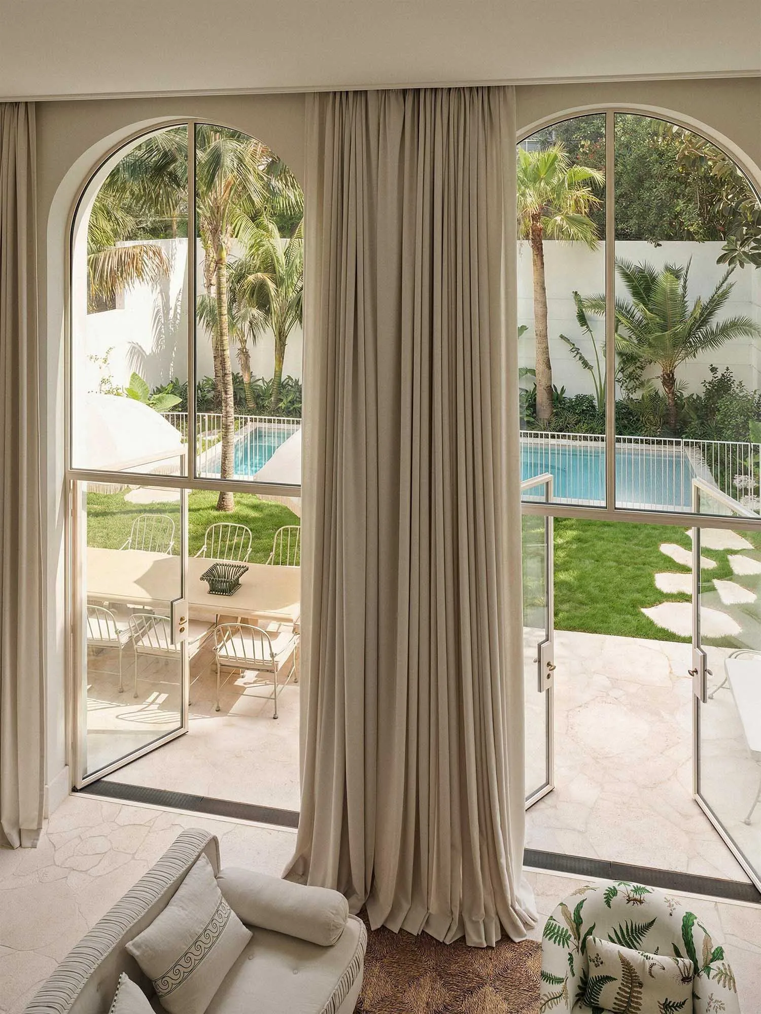 View from a bright sunroom with beige linen curtains, arched glass doors, and outdoor patio furniture overlooking a swimming pool and tropical garden.