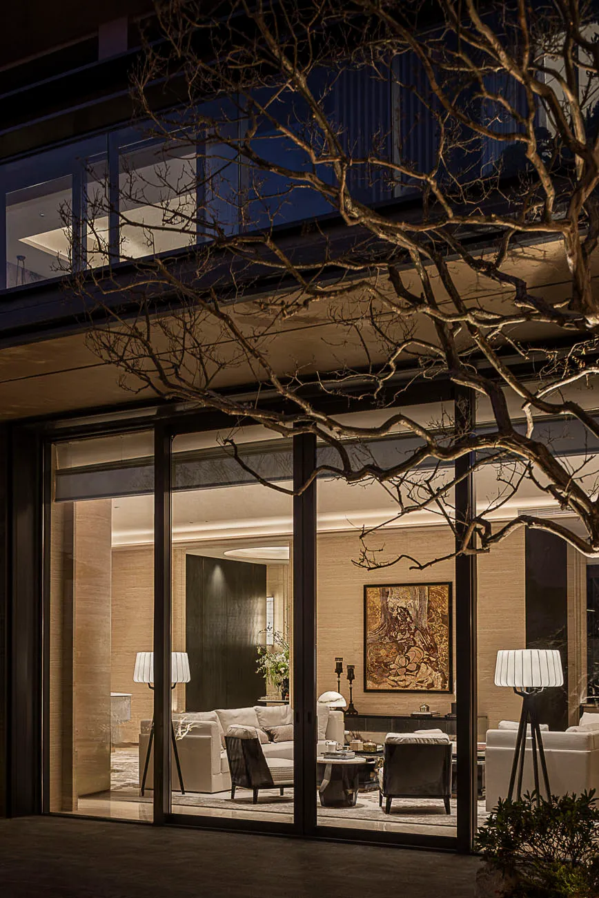 Night view of a modern luxury living room featuring travertine walls, neutral sofas, sculptural floor lamps, and a large artwork, seen through black-framed glass doors with a bare tree in the foreground.