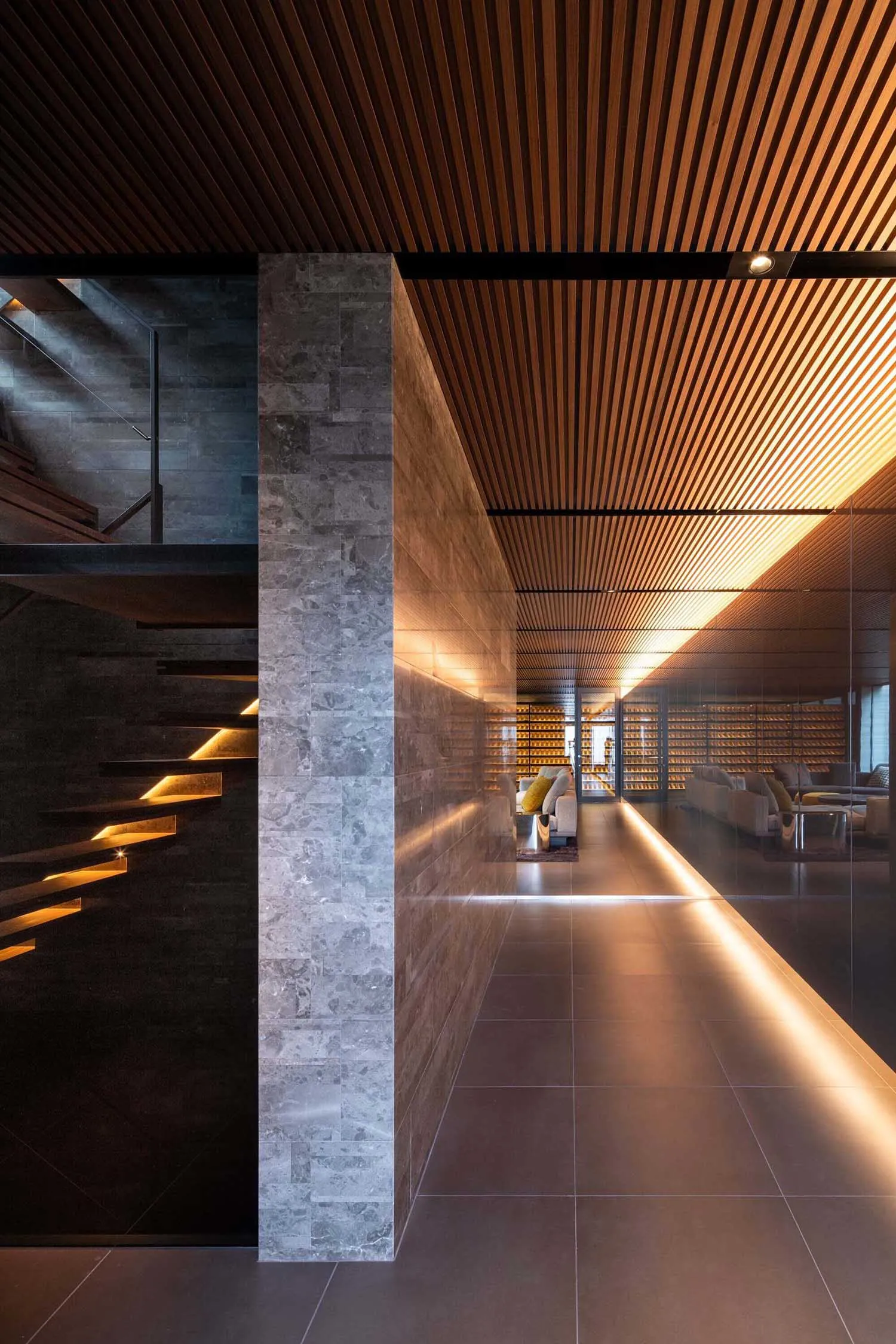 Modern luxury hallway featuring grey marble walls and floors, dark wood slat ceiling with integrated LED lighting, floating staircase with underlit steps, and a view into a living area with wine cellar in the background.