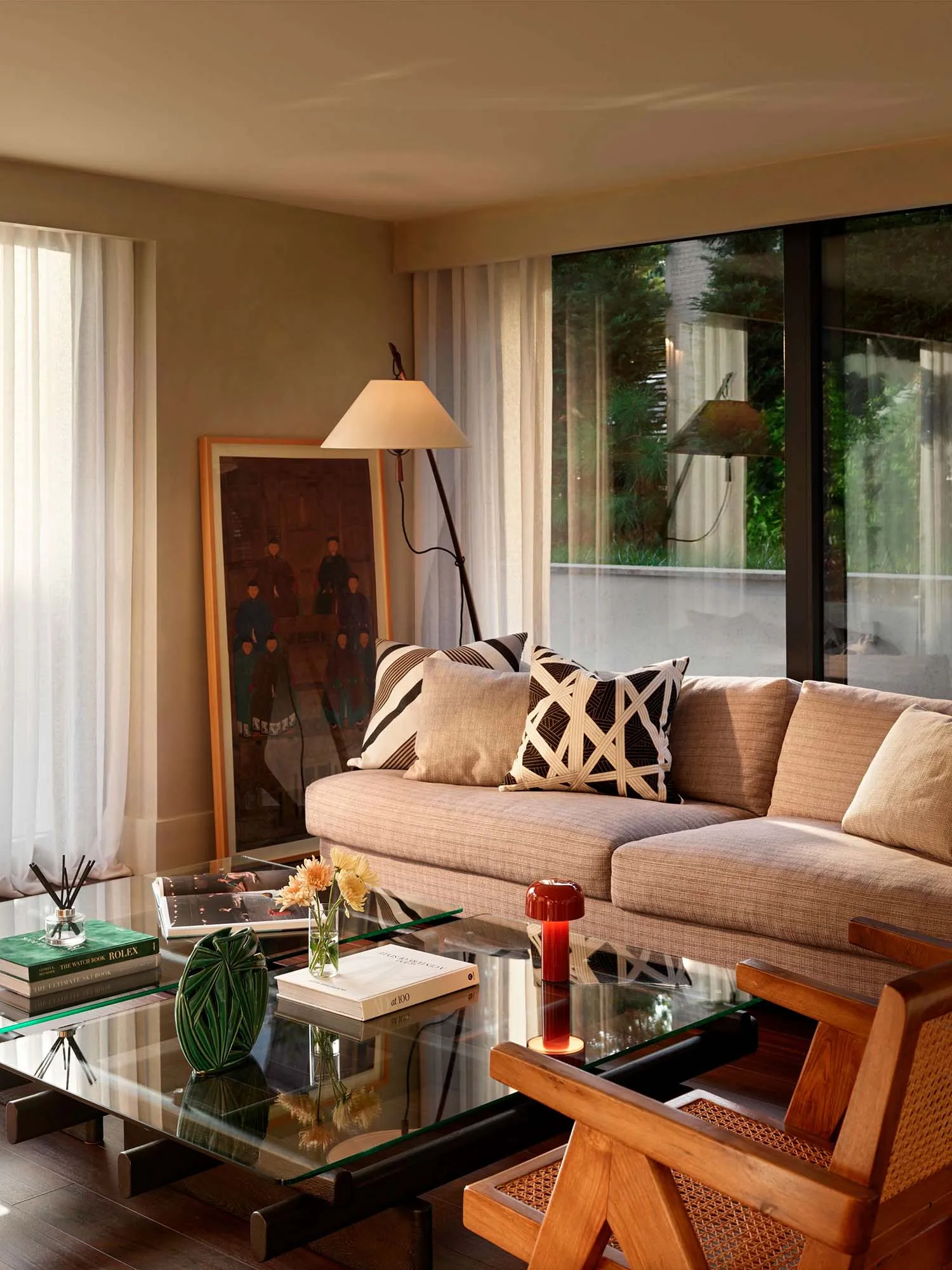 Modern living room featuring a beige linen L-shaped sofa with black and white patterned pillows, a glass coffee table with books and flowers, a wooden armchair with cane detail, and a large framed Asian portrait painting beside a floor lamp.