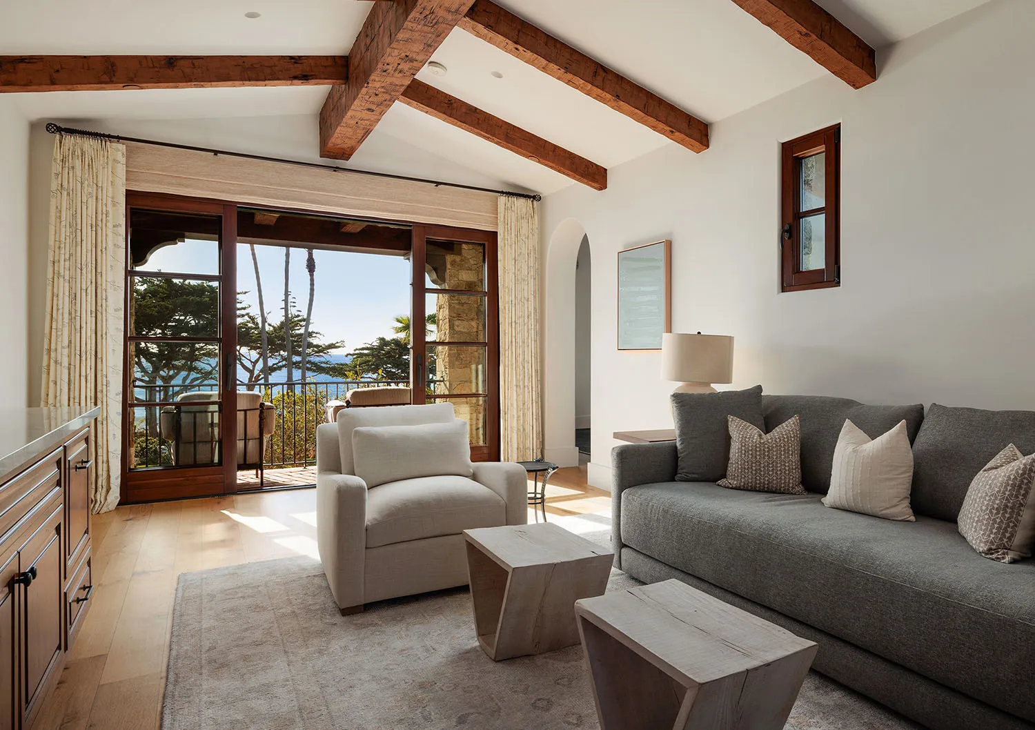 Bright coastal living room with white walls, reclaimed wood ceiling beams, grey sofa, cream armchair, wooden coffee tables, and large glass doors opening to an ocean view with palm trees.