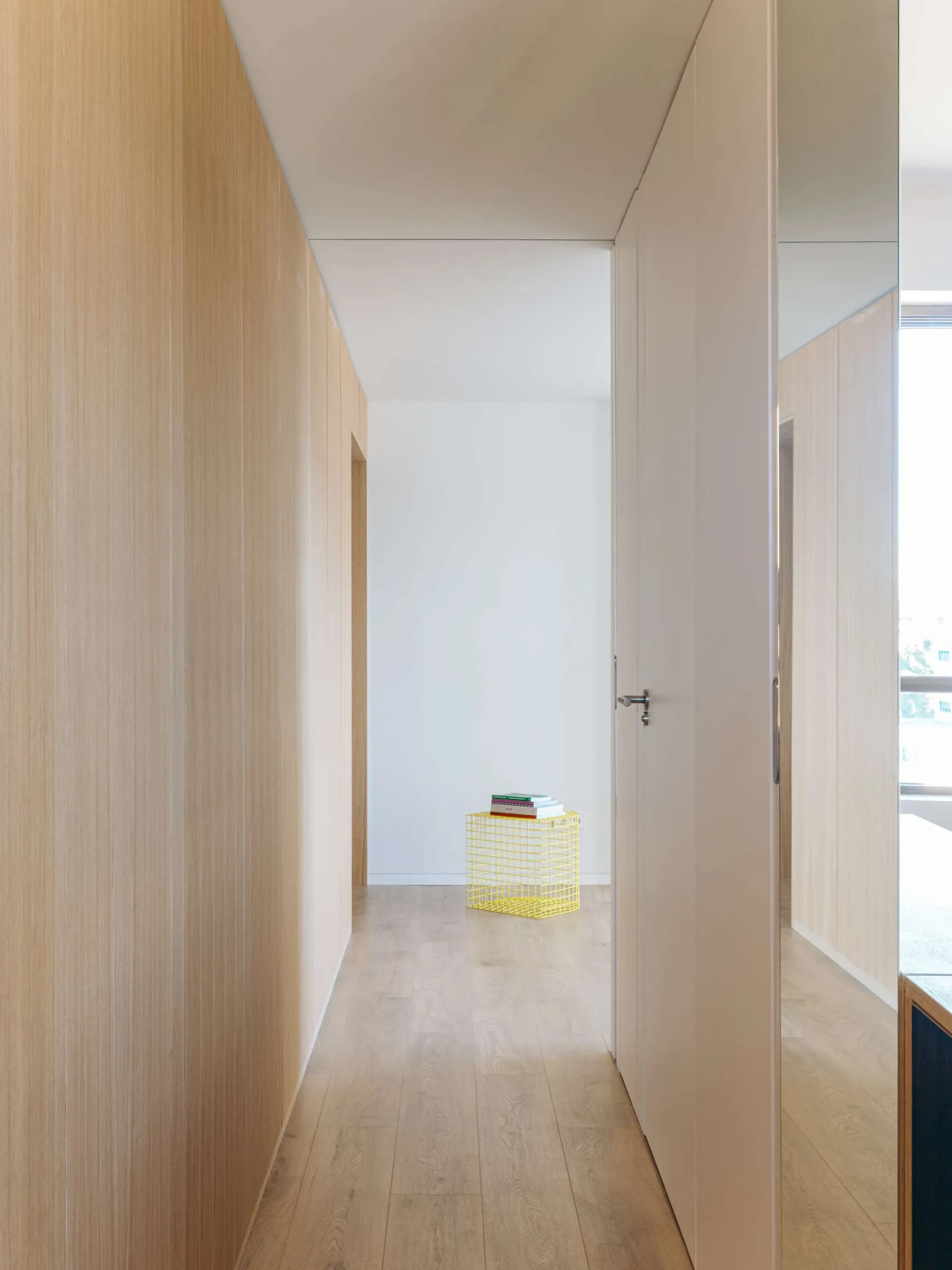 Narrow minimalist hallway lined with light oak wood paneling and cabinetry, featuring a bright yellow wireframe stool holding books at the end of the corridor.