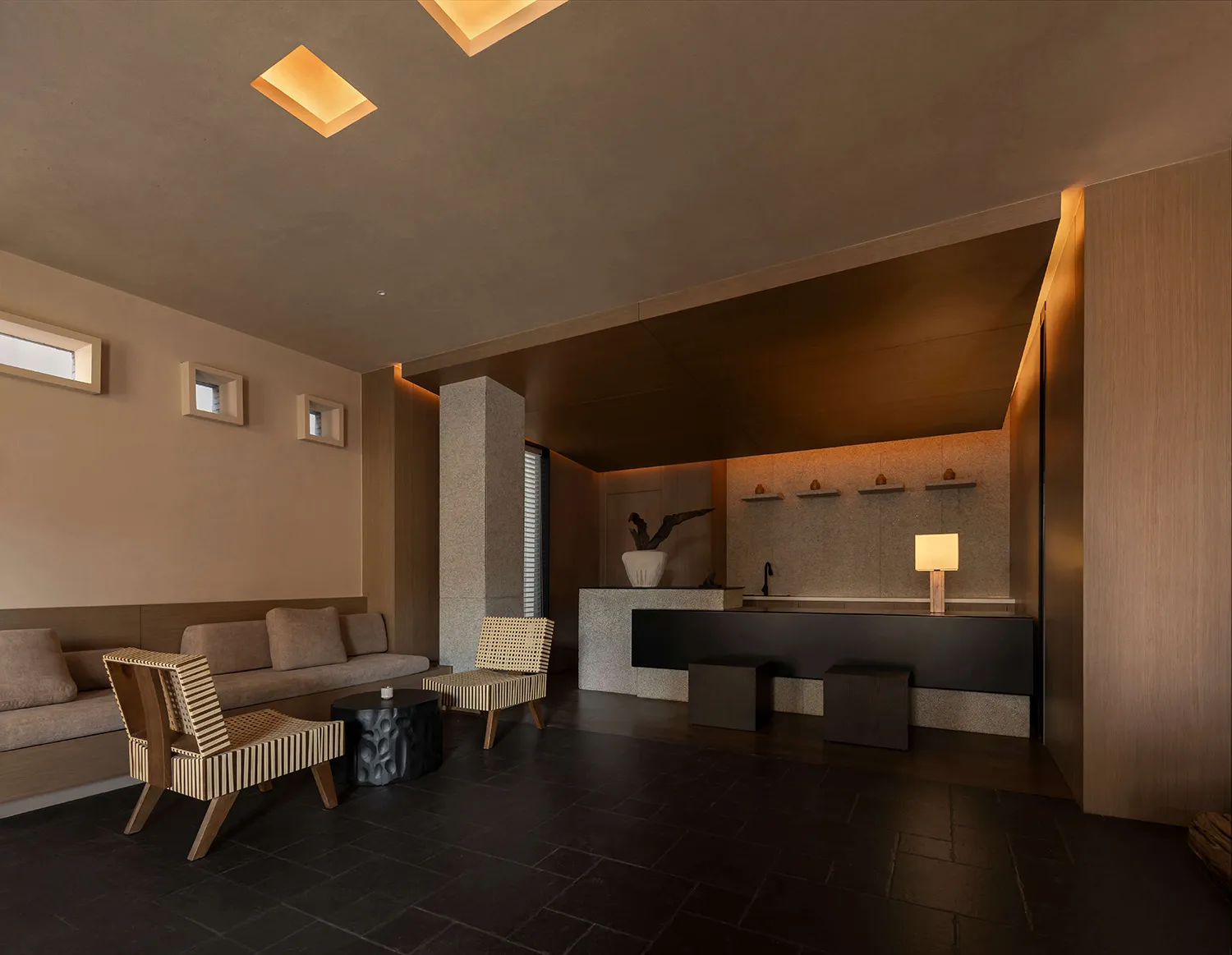 Modern minimalist lounge and reception area featuring exposed concrete walls, dark tiled floor, built-in beige sofa, woven cane armchairs, and a black reception counter with warm recessed lighting.