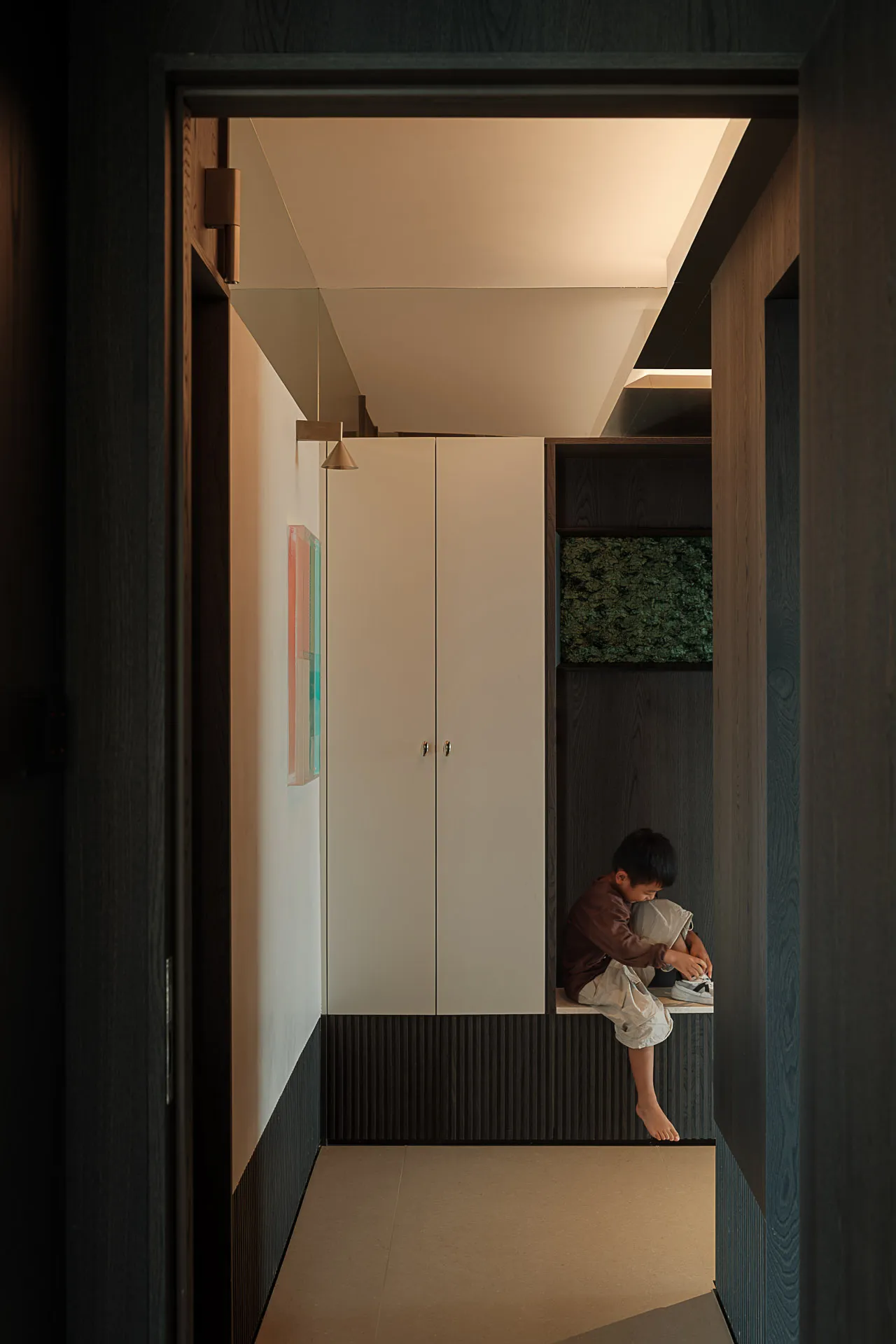 Modern minimalist entryway featuring dark wood paneling, white storage cabinets, a built-in bench, and a young boy putting on shoes under soft overhead lighting.