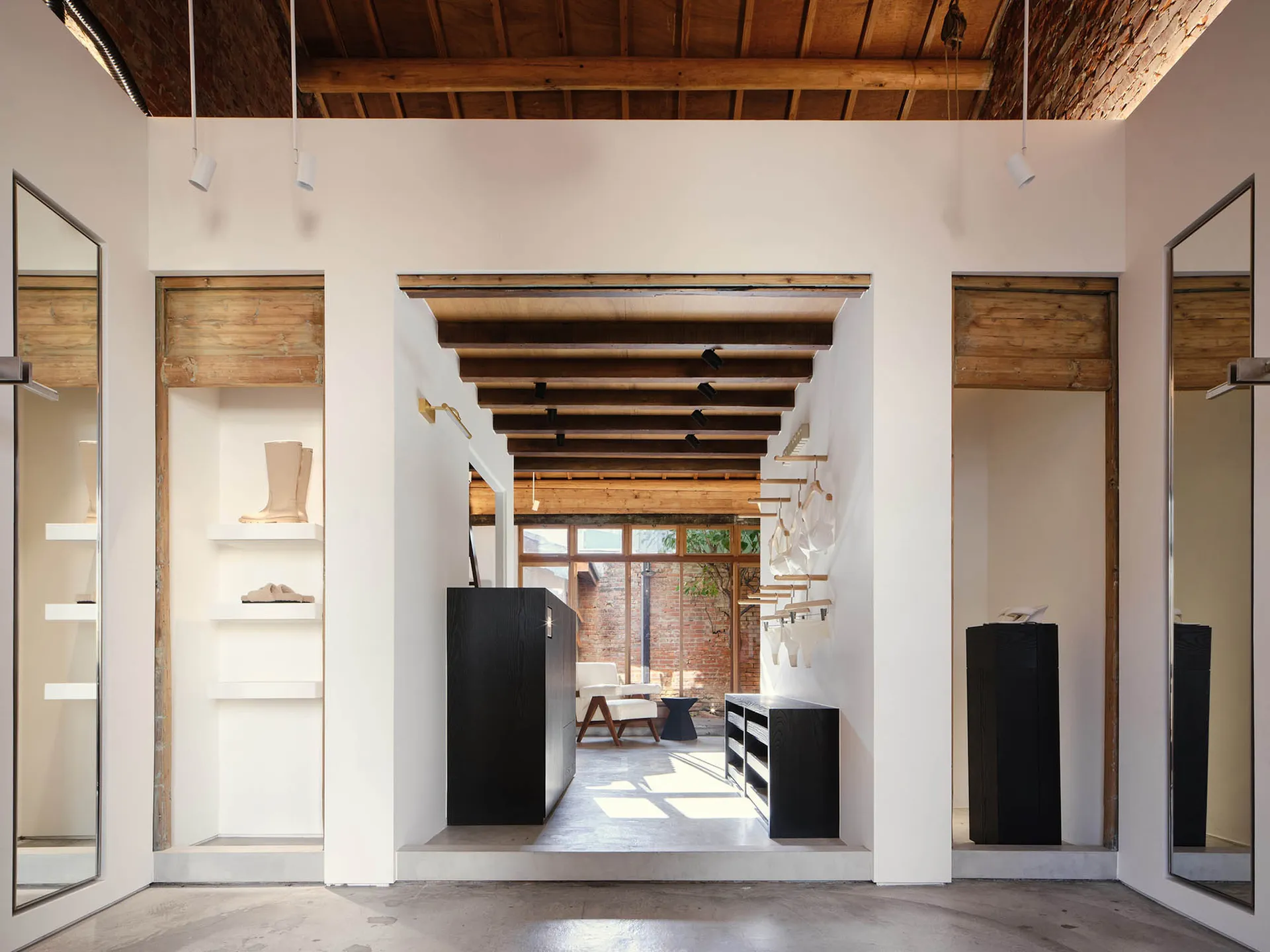Modern minimalist boutique interior featuring white walls, black display fixtures, and prominent reclaimed wooden ceiling beams with natural light from rear courtyard.