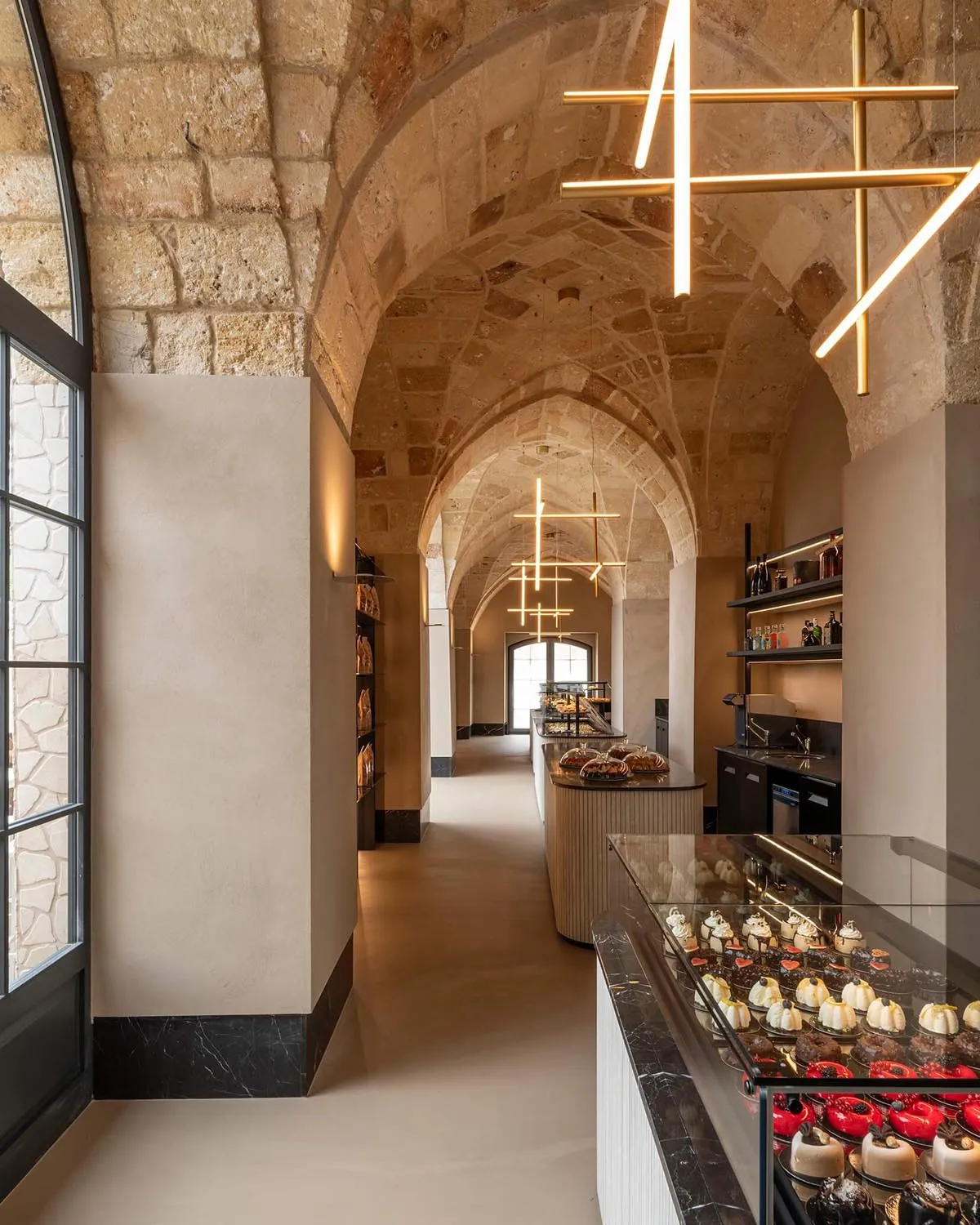 Interior view of a luxurious bakery featuring ancient stone vaulted ceilings, modern linear pendant lights, and glass pastry display cases filled with cakes and desserts.