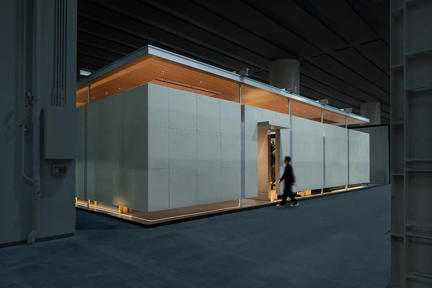 Modern concrete modular pavilion with warm wooden ceiling, integrated LED lighting at the base, and a person walking past to show scale, inside a large industrial hall.