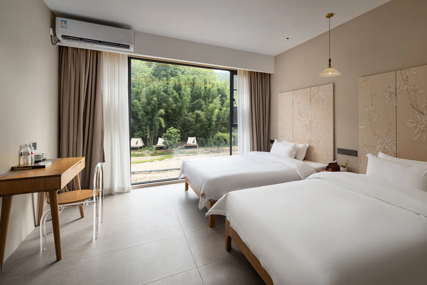 Modern twin bed hotel room with white bedding, floral pattern headboards, wooden desk, and large glass doors opening to a green bamboo forest view.