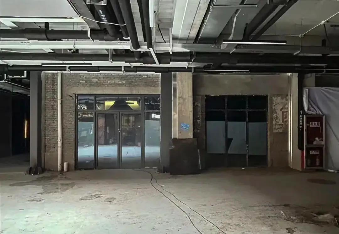 Empty raw industrial loft interior with exposed concrete pillars, brick walls, overhead black piping and ductwork, and large glass doors.