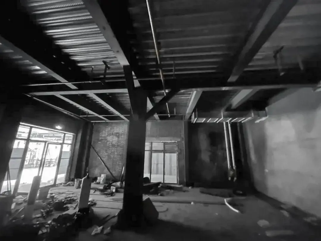 Black and white photograph of an unfinished industrial loft interior showing exposed steel beams, concrete ceiling, structural columns, and large windows with construction debris on the floor.