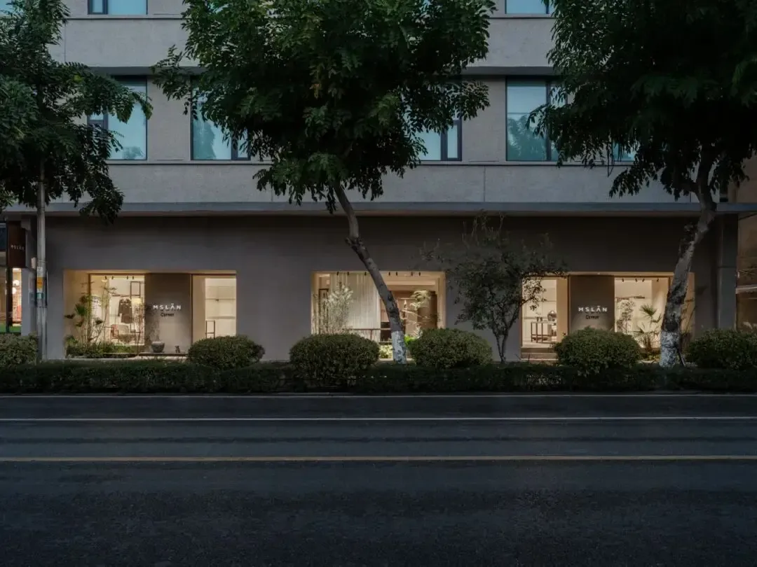 Evening view of a minimalist concrete retail building with warm interior lighting, large glass openings, and trees lining the street.