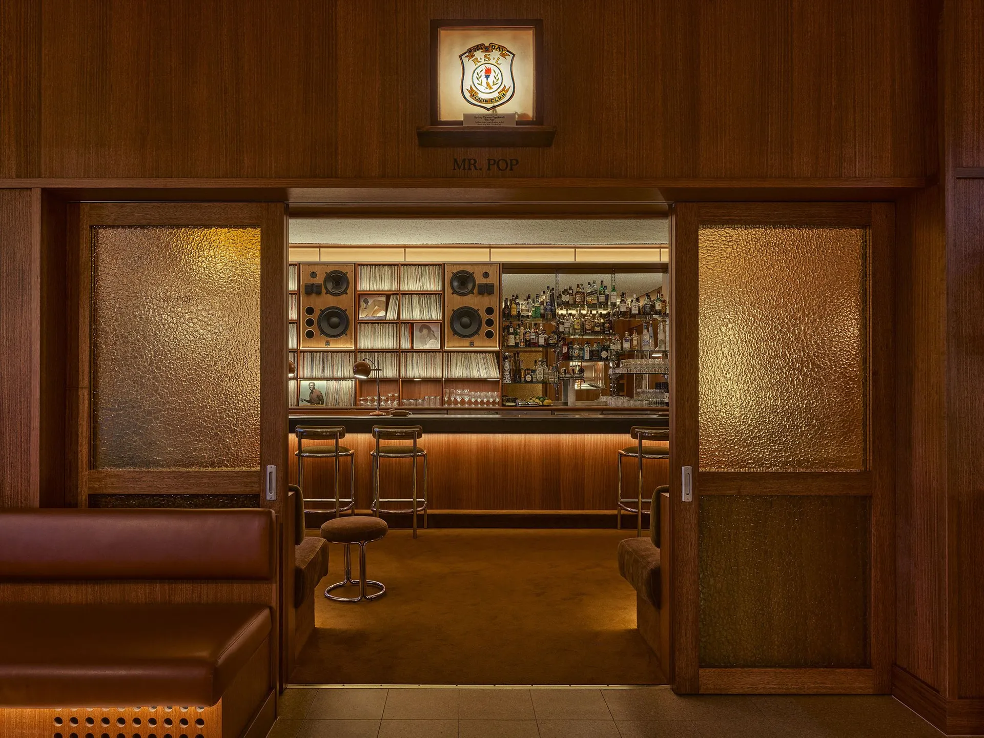 View through textured glass doors into a mid-century modern home bar featuring walnut wood paneling, leather seating, bar stools, vinyl record collection, and backlit bottle display.