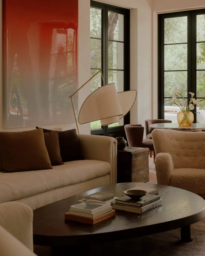 Modern living room featuring a beige boucle sofa with brown pillows, a dark wood oval coffee table stacked with books, a sculptural white and brass floor lamp, a fuzzy boucle armchair, and a large glossy red abstract painting on the wall.
