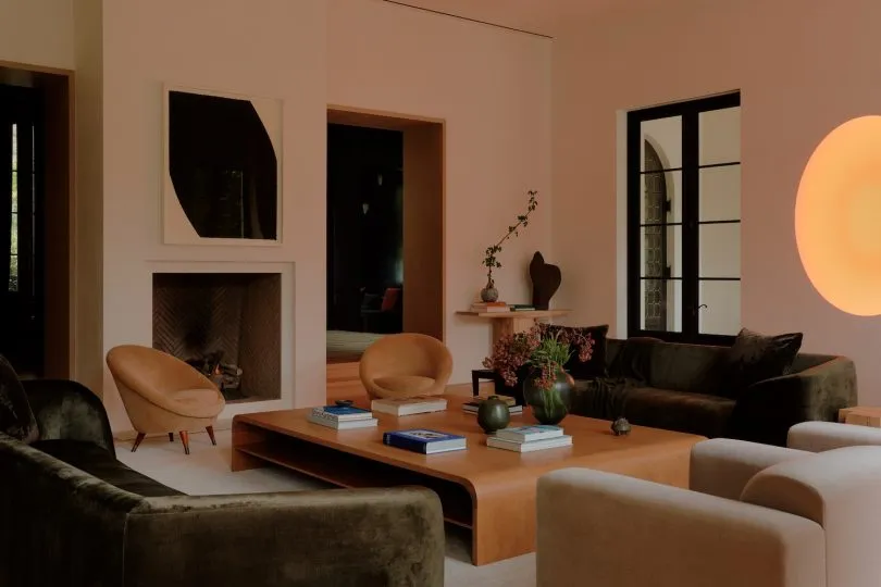 Modern living room featuring dark green velvet sofas, tan curved armchairs, a large oak coffee table, minimalist fireplace, and large spherical wall light.