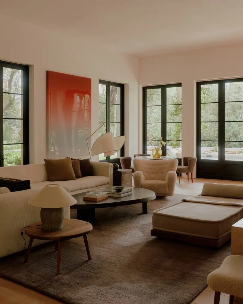 Modern living room featuring a cream boucle sofa, a large red gradient abstract painting, dark-framed glass doors opening to a garden, and layered wooden and boucle seating.