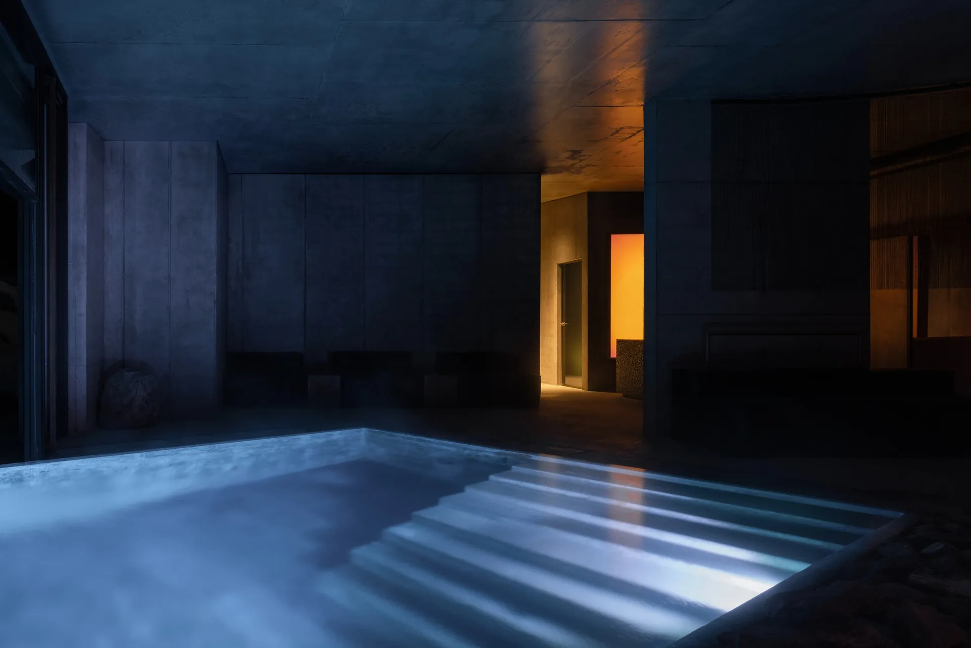 Night view of an indoor swimming pool in a brutalist concrete structure with dramatic blue underwater lighting and warm orange accent lighting in the background corridor.