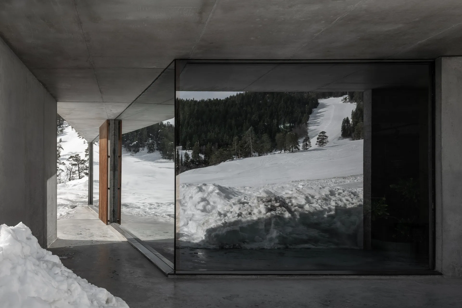 Interior view of a modern concrete mountain house looking out onto a snow-covered alpine forest and ski slope.