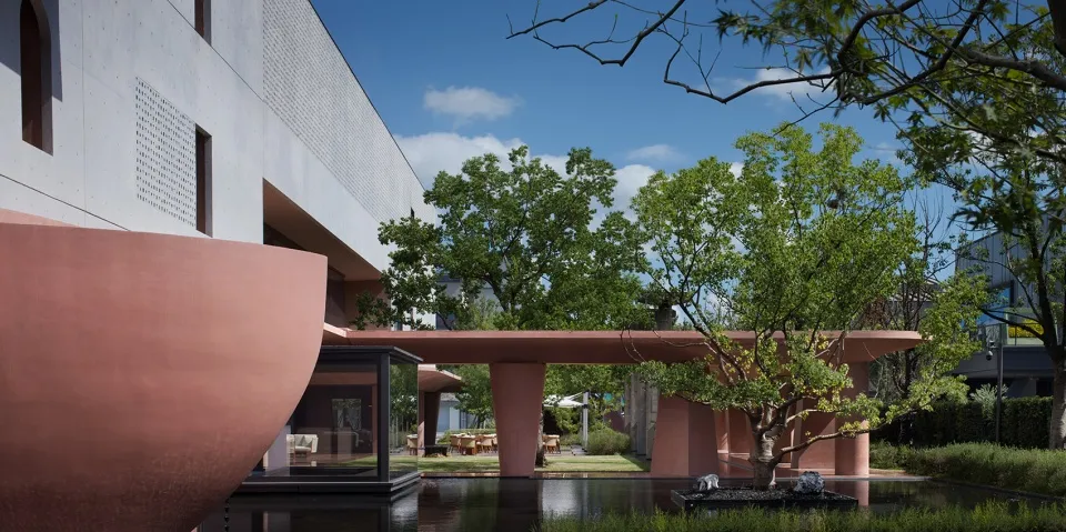 Modern architectural courtyard featuring large terracotta concrete structures, a shallow reflection pool, lush green trees, and a contemporary building with perforated concrete walls.