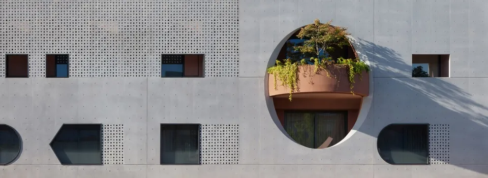 Modern concrete building exterior with geometric windows, perforated panels, and a prominent circular balcony filled with lush greenery and a small tree.