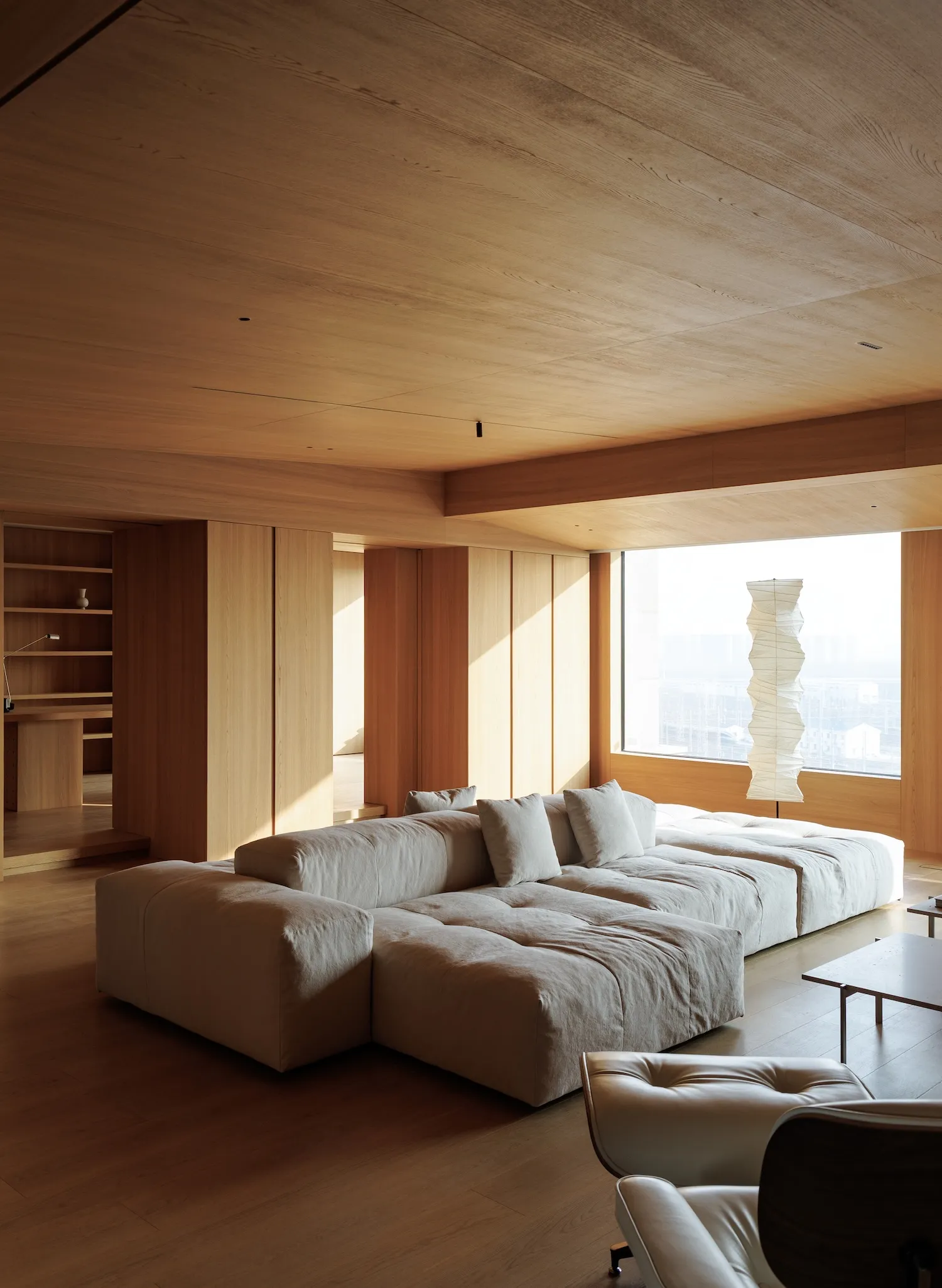 現代 minimalist 客廳,配置大型低矮米白色模組沙發、滿室淺橡木牆面與天花板、紙藝雕塑落地燈,以及前景的皮革休閒椅。