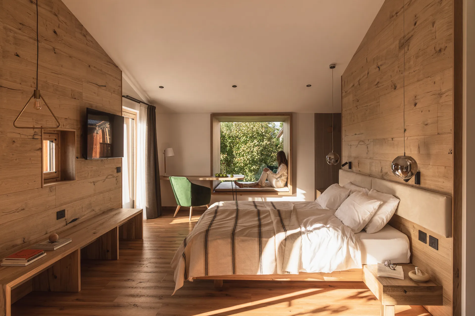 Modern minimalist bedroom with extensive light wood paneling, a large bed with striped linen, built-in window seat with a woman sitting, green velvet chair, and pendant lights.