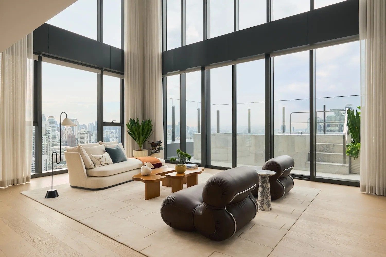 Modern luxury living room featuring a brown leather chaise lounge, cream fabric sofa, wooden coffee tables, and expansive floor-to-ceiling windows overlooking a dense city skyline.