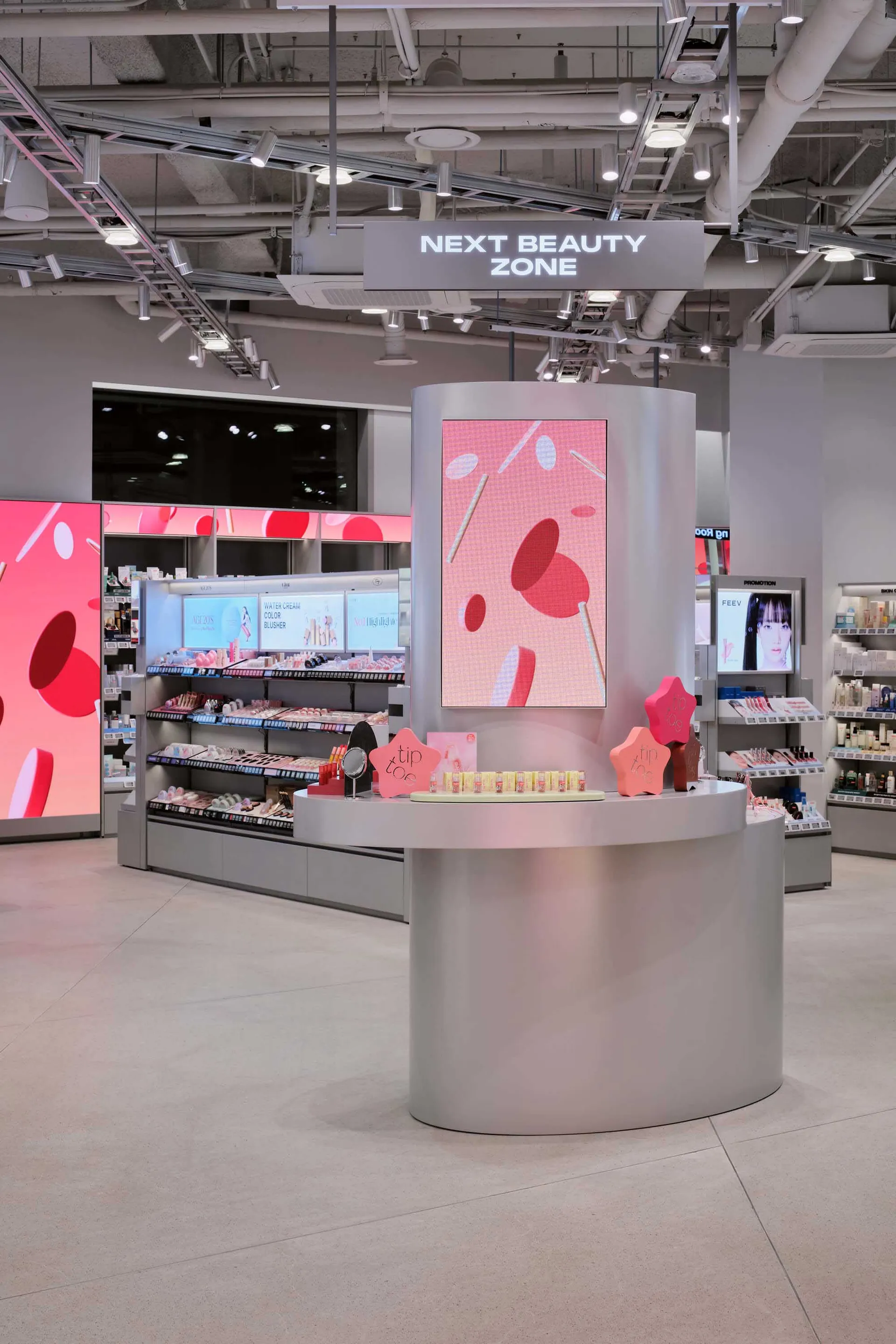 Modern silver curved beauty counter kiosk with large pink digital screen and product displays in a brightly lit retail space