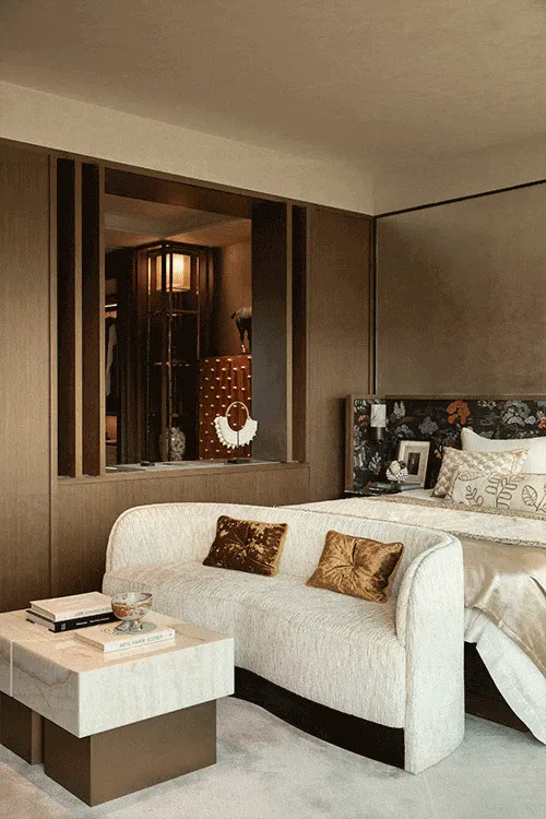 Modern luxury bedroom interior with a curved white bouclé sofa, two gold velvet pillows, marble coffee table with books, walnut wood paneling, and a floral patterned headboard on the bed.