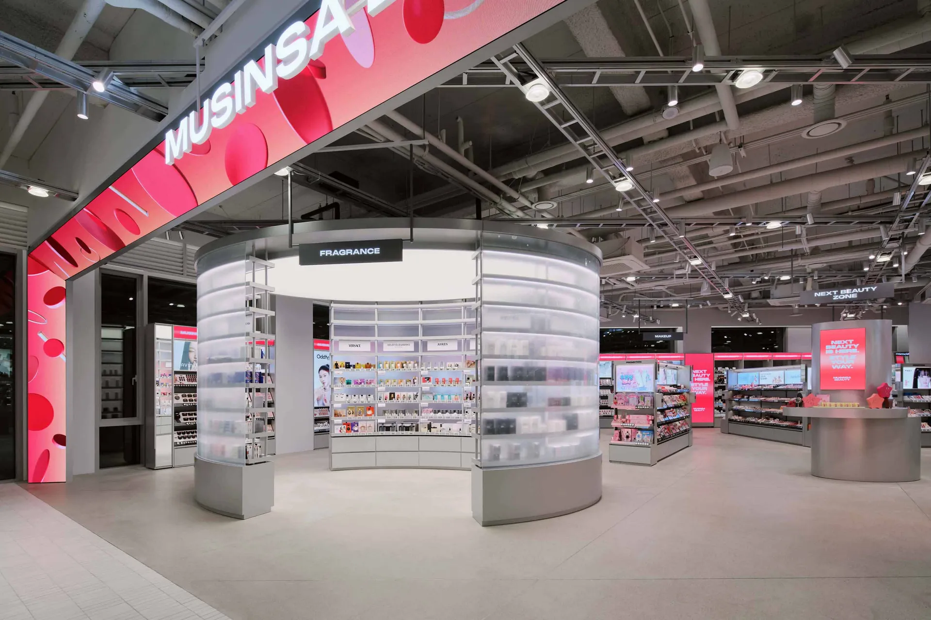 Modern beauty store interior with a large circular fragrance kiosk made of translucent acrylic shelves and integrated lighting, under a pink digital banner reading MUSINSARA.