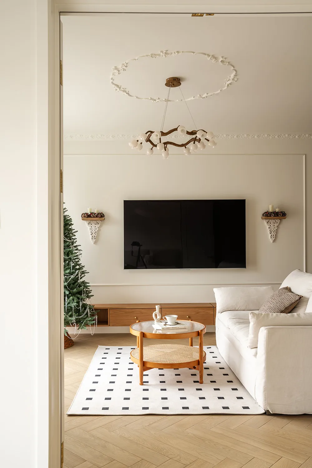 View into a bright living room with white sofa, round oak coffee table on a black and white checkered rug, wall-mounted TV, and a decorated Christmas tree.