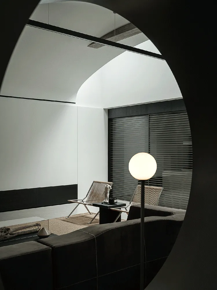 View through a large circular archway into a modern minimalist living room featuring dark leather sofa, two woven rattan lounge chairs, a black side table with sculpture, and a glowing spherical floor lamp.