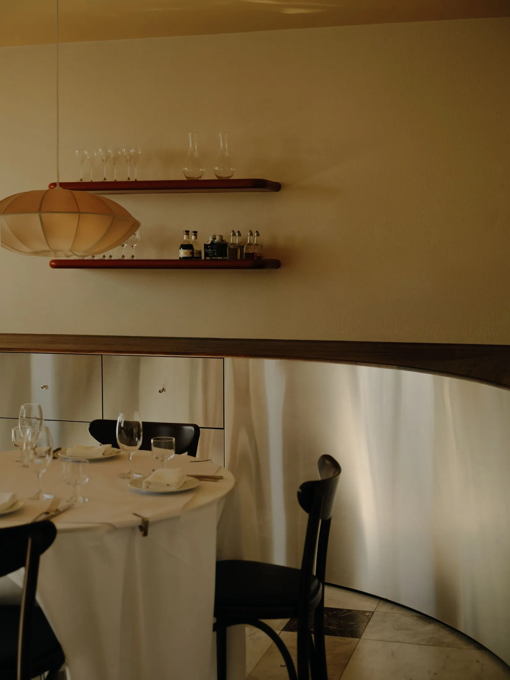 Elegant dining area featuring a curved walnut wall, built-in floating shelves, black dining chairs, and a round table set with white linens under a large pendant lamp.