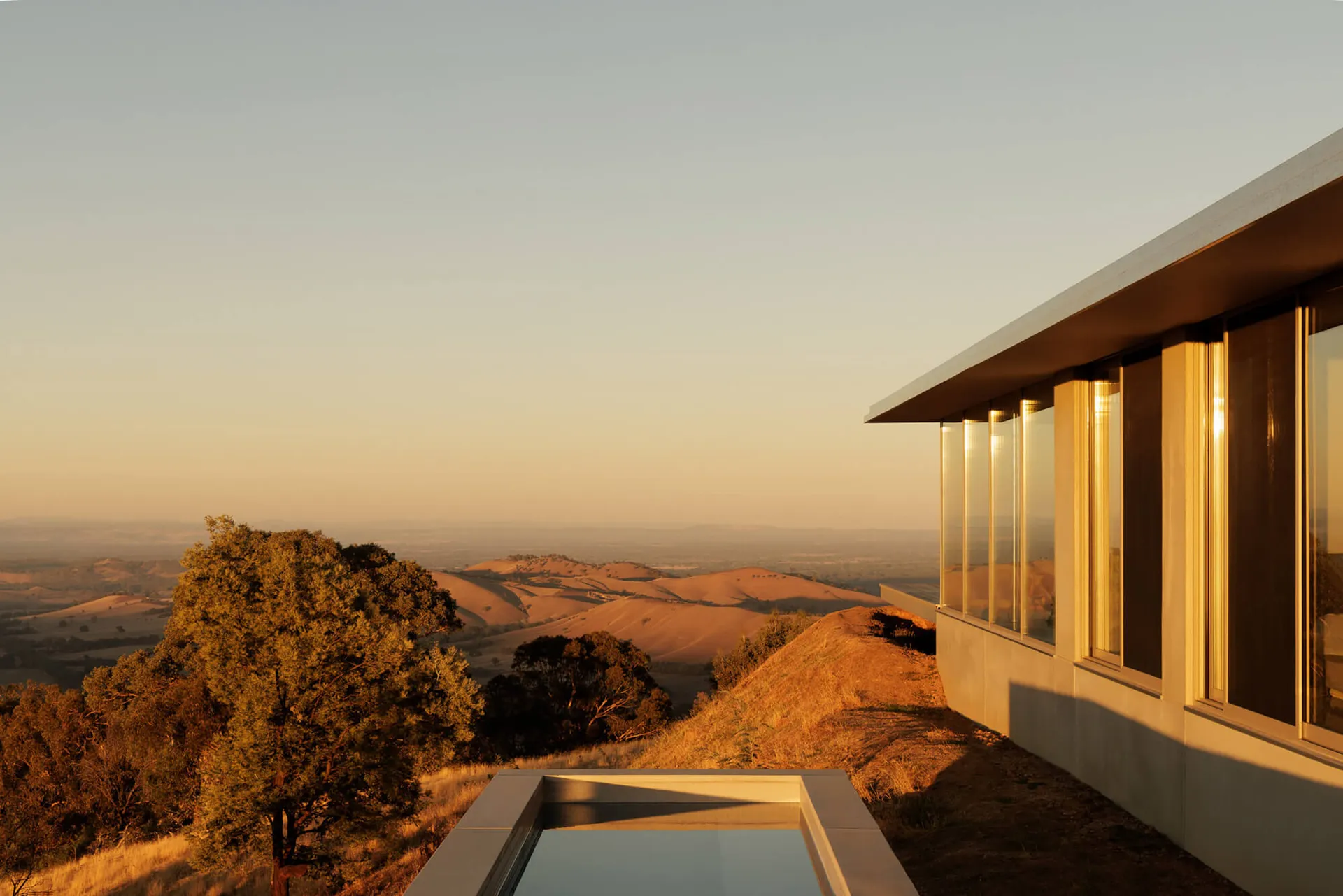 Modern concrete and glass villa with large windows and an infinity pool overlooking a vast golden landscape at sunset.