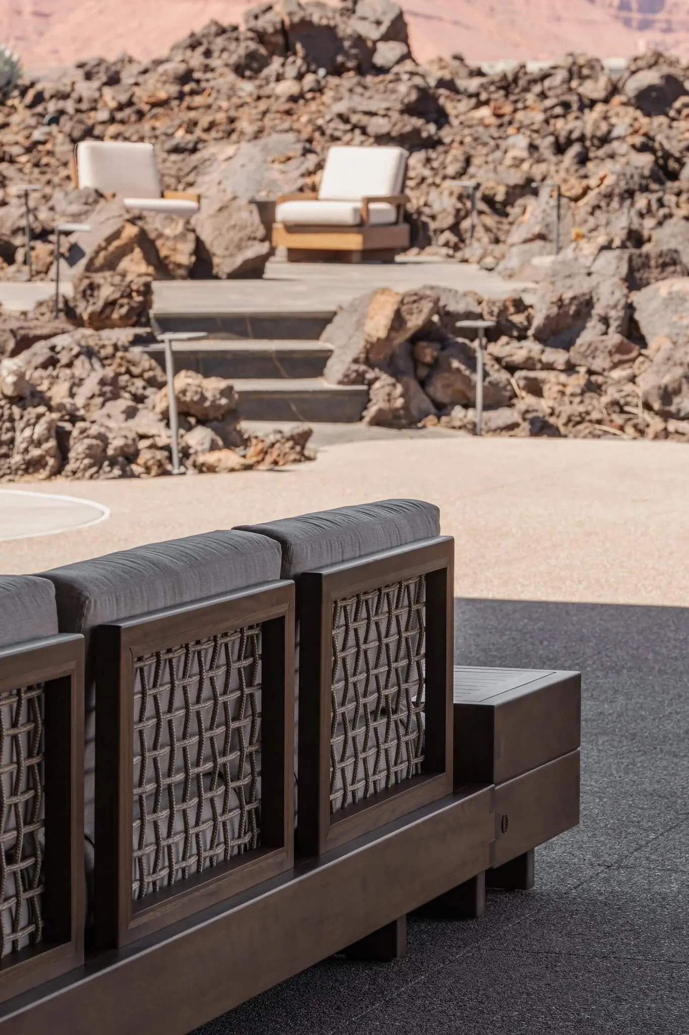 Modern dark teak outdoor sofa with grey cushions and rope detailing positioned on a stone terrace overlooking rugged desert rocks and two background lounge chairs.