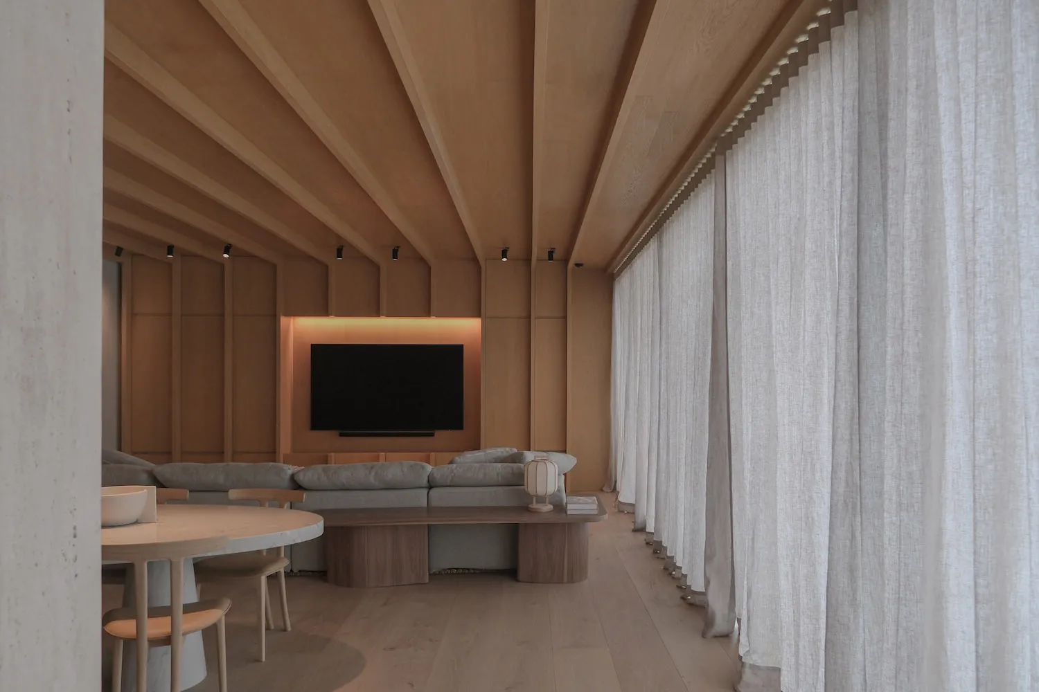 Modern minimalist living room featuring a dramatic sloped oak ceiling, built-in entertainment wall with backlit TV, light grey sectional sofa, round marble dining table, and floor-to-ceiling sheer curtains.