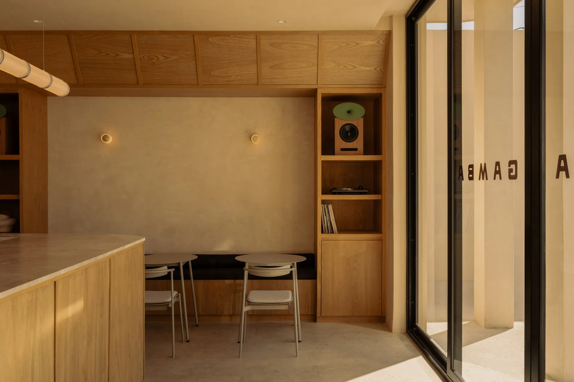 Modern minimalist dining area featuring light oak cabinetry, built-in upholstered bench, round tables with metal chairs, and a tall shelving unit displaying a speaker and vinyl records.