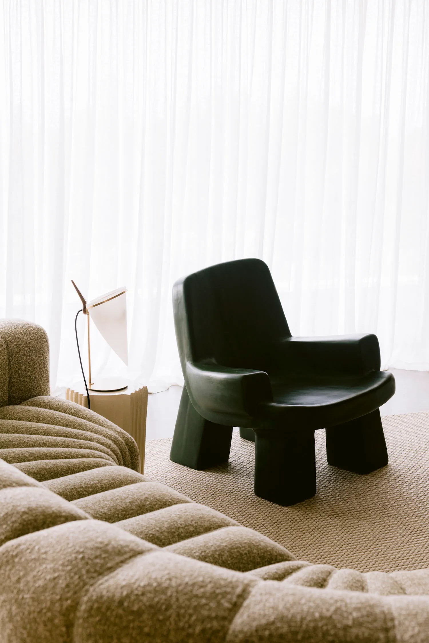 Modern living room corner featuring a black sculptural armchair, a beige boucle sofa, a white table lamp on a fluted side table, and floor-to-ceiling sheer white curtains.