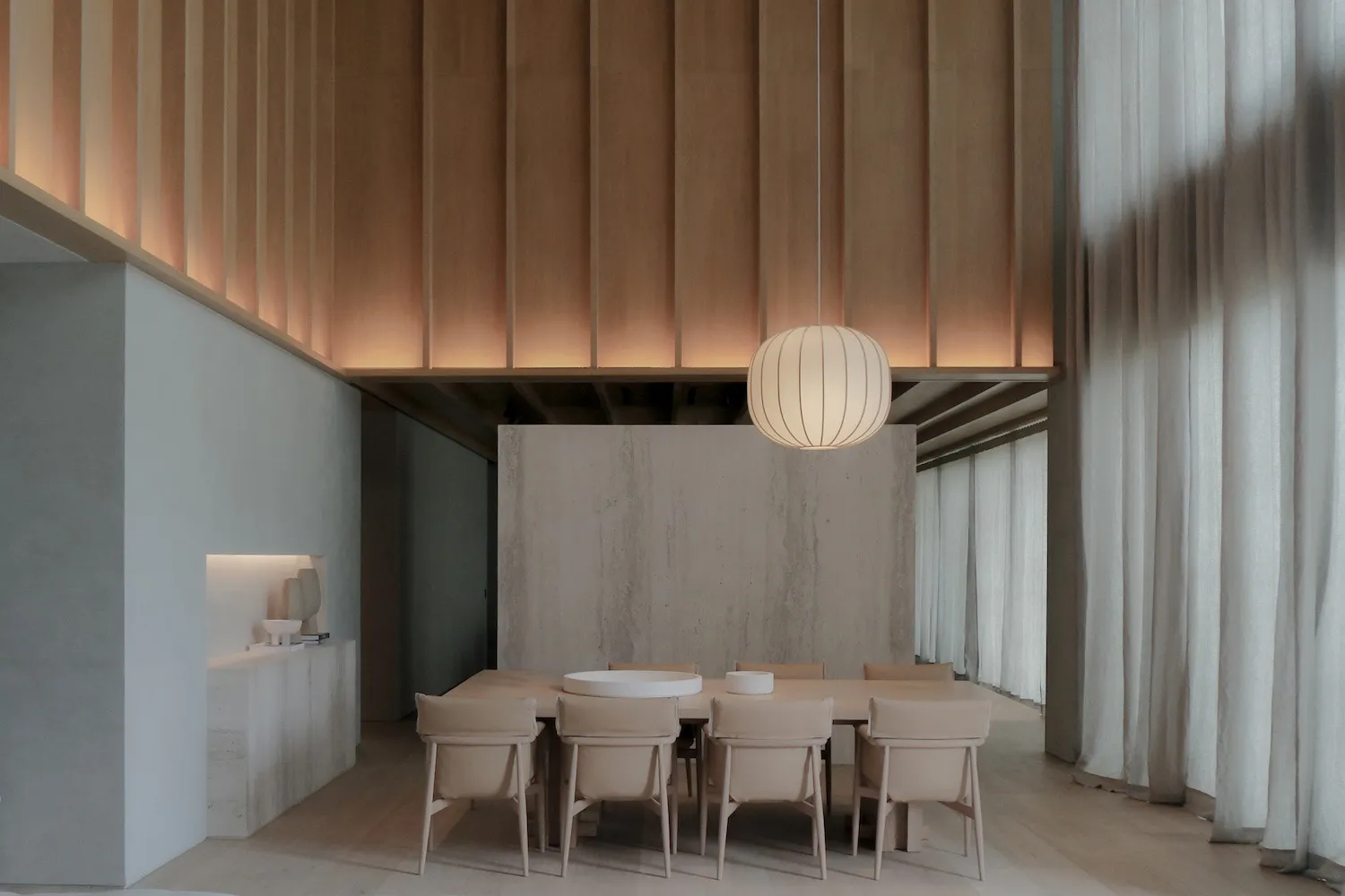 Modern minimalist dining room featuring a long oak dining table with beige upholstered chairs, a large ribbed pendant lamp, vertical wood slat ceiling, and a prominent concrete accent wall.
