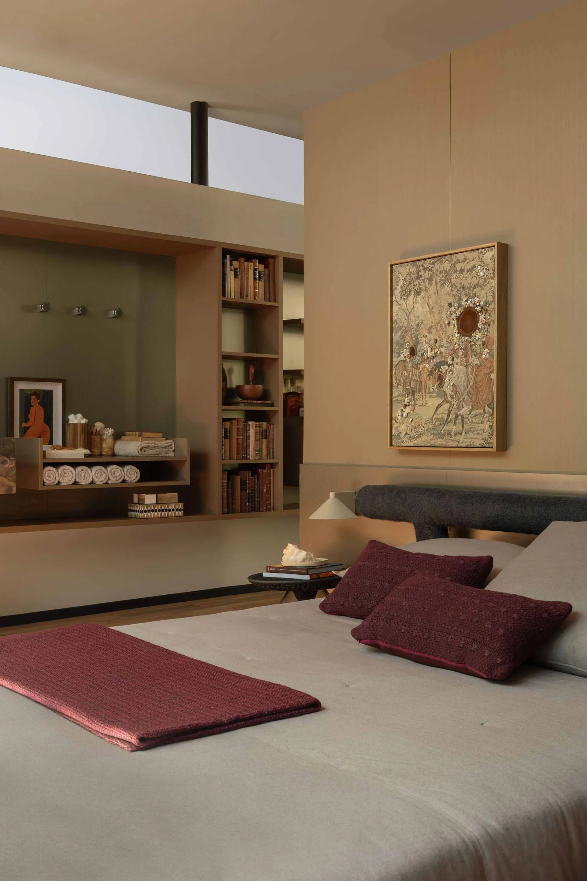 Modern minimalist bedroom with beige walls, built-in wooden shelving, burgundy throw and pillows, and large framed artwork above the bed.