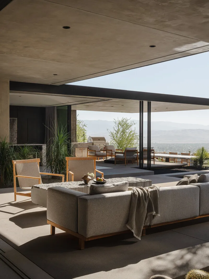 Modern outdoor living area with concrete ceiling, wooden-framed sofas, woven lounge chairs, and panoramic desert mountain views.