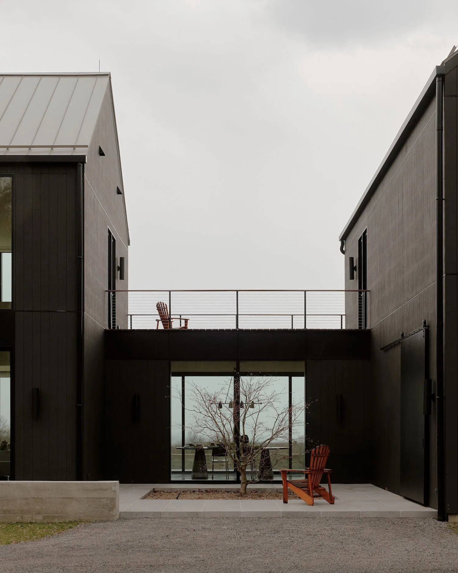 Modern black charred wood house exterior featuring a central courtyard with large glass windows, a small tree, and two wooden Adirondack chairs on an upper balcony and ground level.