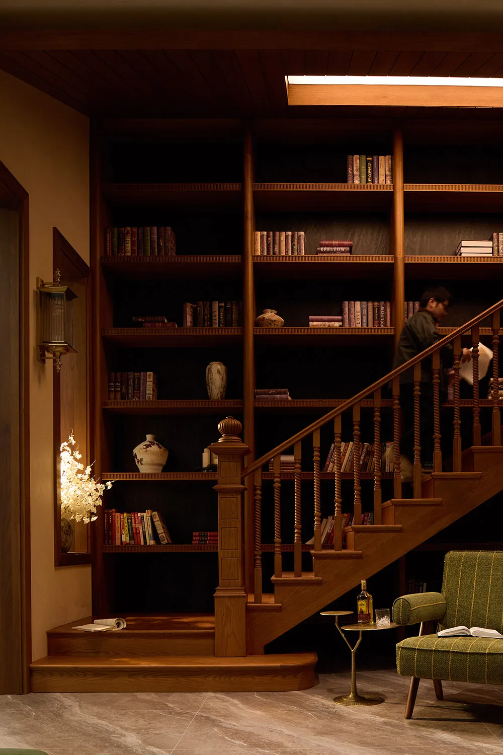 Warmly lit interior featuring a walnut staircase with integrated bookshelves, a green striped armchair, and a person browsing books on the upper level.